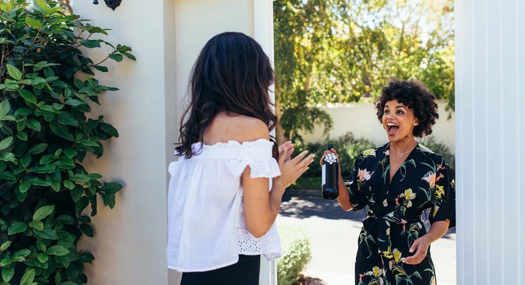 Excited african woman attending a female friend's housewarming party. Woman giving a wine bottle her friends at entrance door.